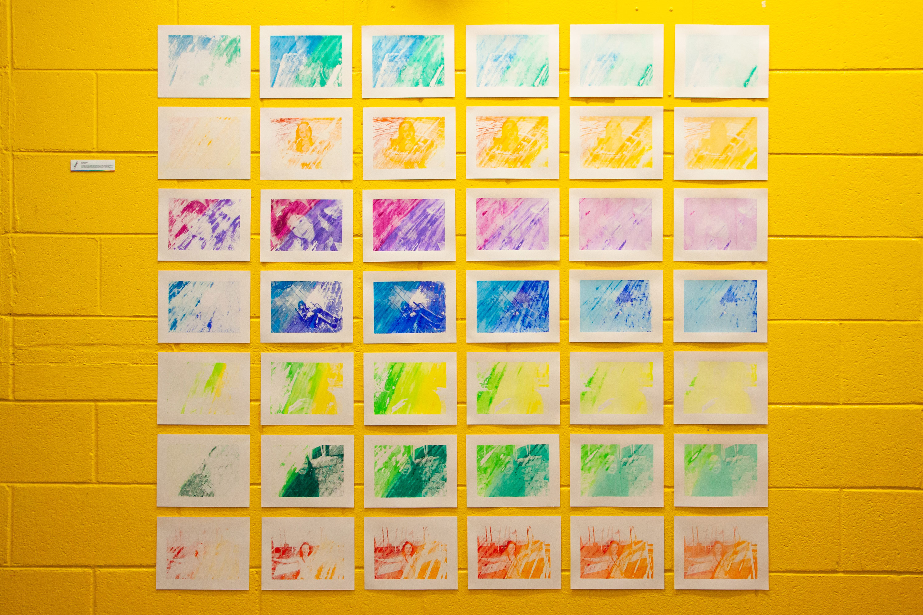 An installation of 42 colorful monoprints, organized into a grid of 6 columns and 7 rows. Down each column are prints of various photographs in multi-colored gradients, arranged in chronological order. Across each row is a series of ghost prints created from the prints on the left. The prints are installed on a yellow wall with a small label next to the grid.