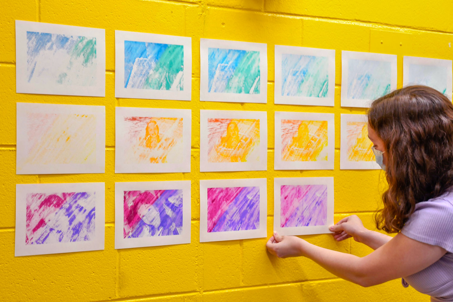 Elora installing the prints onto a yellow wall, arranged in a grid. The photo is taken from the back side. She has brown curly hair and is wearing a mask and a light purple shirt. Only the side of her face is visible, and she is using her hands to apply a print to the 3rd row.