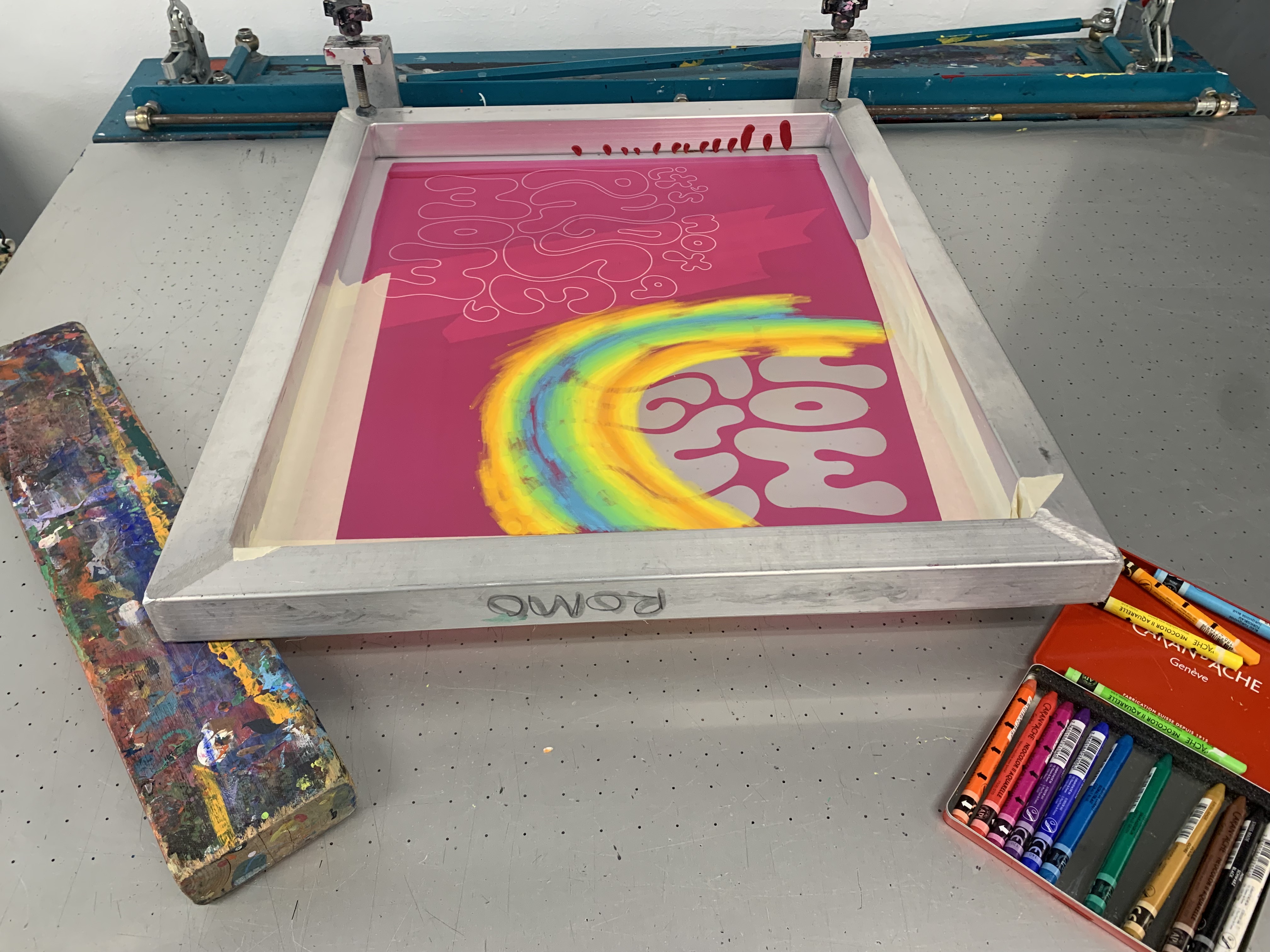 A print table set up for mono-screen printing, with a screen propped up on a colorful ink-covered wooden block. On the screen, there is pink emulsion with a stencil that has been exposed to read 'it's not a phase, mom' in bubbly letters. In this image, the type isn't readable because it is colored over partly with a gradient of orange, yellow, green, and blue pastel. To the right of the screen, there is an open red case of water-soluble pastels in all colors.